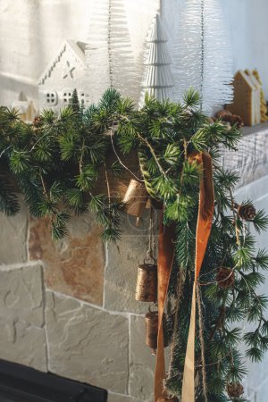 Foto de Modernos árboles de Navidad, casas, conos de pino y ramas de abeto en la chimenea mantel con campanas de latón y cinta. Elegante chimenea de Navidad decorada en la sala de estar escandinava mínima - Imagen libre de derechos