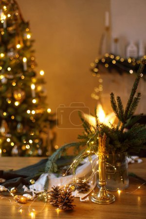 Téléchargez les photos : Bougie élégante, lumières dorées, cônes de pin et ornements sur table en bois contre arbre de Noël décoré élégant et cheminée avec éclairage festif. Vacances d'hiver dans l'atmosphère à la maison de fête - en image libre de droit
