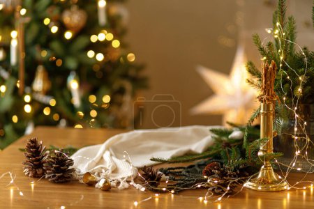 Foto de Vela con estilo, luces doradas, conos de pino y adornos en la mesa de madera contra el elegante árbol de Navidad decorado con iluminación festiva. Vacaciones de invierno atmosféricas en el hogar rústico festivo - Imagen libre de derechos