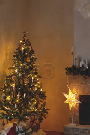 Foto de Elegante sala de estar de Navidad con moderno árbol de Navidad decorado y decoración festiva en la chimenea mantel con luz dorada. Nochebuena atmosférica en la sala escandinava. Felices Fiestas! - Imagen libre de derechos