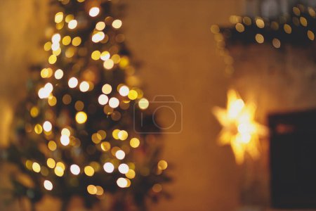 Foto de Habitación de Navidad bokeh dorado. Imagen borrosa de la víspera de Navidad atmosférica en la chimenea. Árbol de Navidad iluminado desenfocado y chimenea festiva con luces en la noche. - Imagen libre de derechos