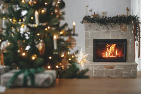 Foto de ¡Feliz Navidad! Elegante chimenea decorada con chimenea festiva, árbol decorado con luces doradas y regalo en la mesa. Nochebuena atmosférica en la chimenea, espacio para el texto - Imagen libre de derechos