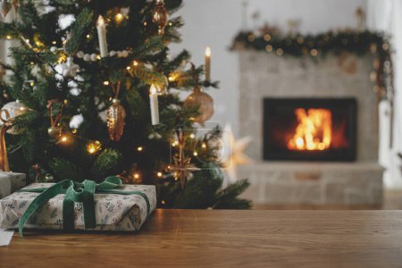 Téléchargez les photos : Élégant cadeau de Noël à proximité contre l'arbre décoré et cheminée en feu. Joyeux Noël ! Beau cadeau enveloppé moderne avec ruban vert sur table en bois. Fond de Noël, espace de copie - en image libre de droit