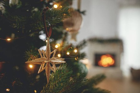 Foto de ¡Feliz Navidad! Elegante estrella de oro de Navidad en el árbol de cerca contra la chimenea en llamas. Hermoso árbol de Navidad decorado con adornos vintage, cintas y luces. Fondo de Navidad. - Imagen libre de derechos