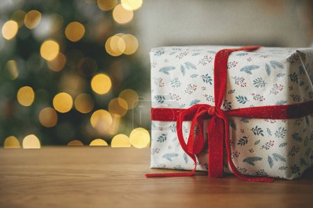 Photo for Stylish christmas gifts close up against tree lights bokeh. Merry Christmas! Beautiful modern wrapped present with red ribbon on wooden table. Christmas background, copy space - Royalty Free Image