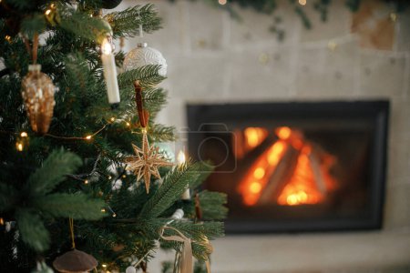 Photo for Stylish christmas tree against burning fireplace. Beautiful decorated christmas tree with vintage baubles and ribbons on background of fireplace. Atmospheric christmas eve. Merry Christmas! - Royalty Free Image