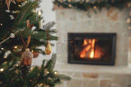 Foto de Elegante bellota de oro de Navidad en el árbol de cerca contra la chimenea en llamas. Hermoso árbol de Navidad decorado con adornos vintage, cintas y luces. Fondo de Navidad. Feliz Navidad.! - Imagen libre de derechos