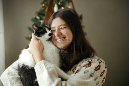 Foto de Mujer feliz en suéter acogedor abrazando gato lindo en el fondo del árbol de Navidad decorado con estilo en la habitación festiva. Propietario con mascotas y vacaciones de invierno. Feliz Navidad.! - Imagen libre de derechos