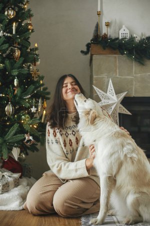 Téléchargez les photos : Femme heureuse dans un pull confortable étreignant et jouant avec un chien blanc mignon sur fond d'arbre de Noël élégant dans une pièce festive. Vacances d'hiver et animaux domestiques. Joyeux Noël! - en image libre de droit