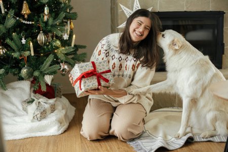 Foto de ¡Feliz Navidad! Mujer en suéter acogedor regalo de Navidad de apertura con lindo perro blanco en el fondo del árbol de Navidad con estilo. Vacaciones de invierno. Propietario con mascota celebrando en sala festiva - Imagen libre de derechos