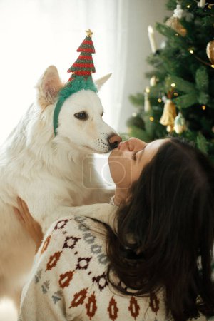 Foto de Mujer feliz en suéter acogedor abrazando lindo perro blanco en accesorio festivo en el fondo del elegante árbol de Navidad. Vacaciones de invierno. Propietario y mascota jugando en la habitación festiva. Feliz Navidad.! - Imagen libre de derechos