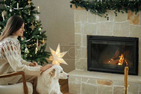 Foto de Mujer en suéter acogedor acariciando lindo perro blanco en el fondo del elegante árbol de Navidad y chimenea caliente. Vacaciones de invierno. Propietario con mascota relajante en habitación festiva. Feliz Navidad.! - Imagen libre de derechos