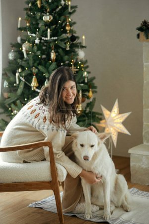 Téléchargez les photos : Femme en pull confortable caressant chien blanc mignon sur fond d'arbre de Noël élégant avec des lumières dorées.Vacances d'hiver. Propriétaire avec animal de compagnie relaxant dans une chambre festive. Joyeux Noël! - en image libre de droit