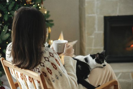 Téléchargez les photos : Hygge vacances d'hiver. Femme en pull confortable assis avec chat mignon et boire du thé chaud sur fond de cheminée et arbre de Noël élégant. Propriétaire avec animal de compagnie relaxant. Joyeux Noël! - en image libre de droit