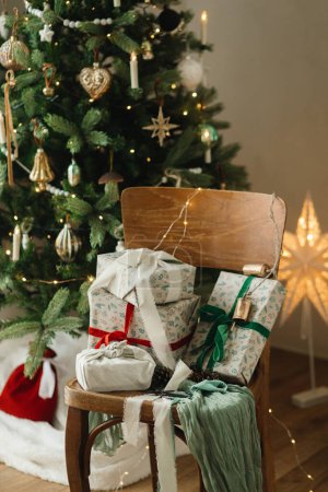 Téléchargez les photos : Cadeaux de Noël enveloppés élégants avec ruban sur une vieille chaise en bois sur fond d'arbre de Noël décoré avec des boules vintage et des lumières festives. Joyeux Noël et Joyeuses Fêtes! - en image libre de droit