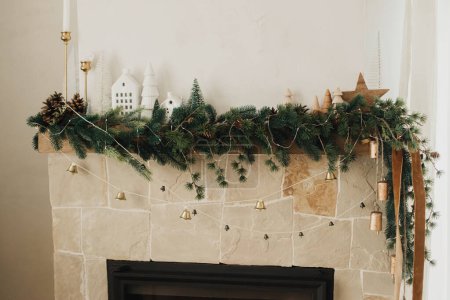 Foto de Elegante chimenea mantel decorado con ramas de Navidad, guirnalda campanas, adornos de madera y decoración de la casa. Chimenea de Navidad rústica festiva en la moderna casa de campo sala de estar - Imagen libre de derechos