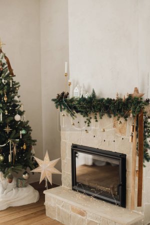 Foto de Elegante chimenea decorada con ramas de Navidad, decoraciones y campanas en el fondo del moderno árbol de Navidad con luces. Sala de estar de Navidad rústica en casa de campo moderna - Imagen libre de derechos