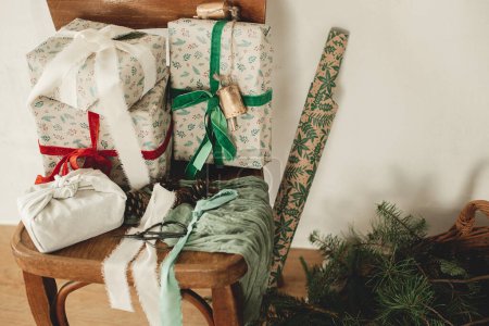 Foto de Feliz Navidad y Felices Fiestas! Elegantes regalos de Navidad envueltos con cintas en una vieja silla de madera, papel festivo y canasta de mimbre con ramas de abeto en la habitación rural. Regalos ecológicos - Imagen libre de derechos
