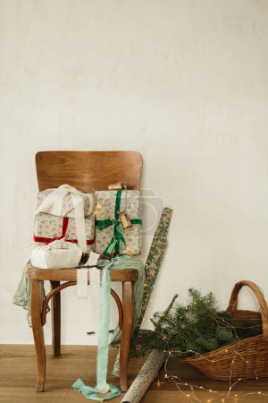 Photo for Stylish wrapped christmas gifts with ribbons on old wooden chair, festive paper and wicker basket with spruce branches in rural room, rustic still life. Merry Christmas and Happy Holidays! - Royalty Free Image
