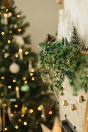 Téléchargez les photos : Manteau de cheminée élégant décoré avec des branches de Noël, guirlande de cloches, ornements sur fond de lumières de sapin de Noël. Festive décor de cheminée rustique dans le salon de ferme moderne - en image libre de droit