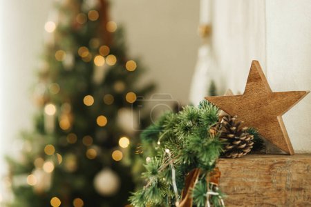 Foto de Elegante estrella de madera de Navidad, ramas de abeto en la chimenea mantel de cerca en el fondo de las luces del árbol de Navidad. Decoración festiva chimenea rústica en la moderna casa de campo sala de estar - Imagen libre de derechos