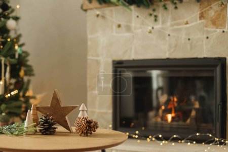 Photo for Stylish christmas wooden trees, star, pine cones and fir branches on table against burning fireplace. Modern rustic eco friendly decor for winter holidays in farmhouse living room - Royalty Free Image
