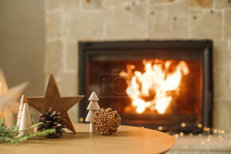 Téléchargez les photos : Élégants arbres de Noël en bois, étoiles, cônes de pin et branches de sapin sur la table contre la cheminée en feu. Moderne décor rustique écologique pour des vacances d'hiver dans le salon de la ferme - en image libre de droit