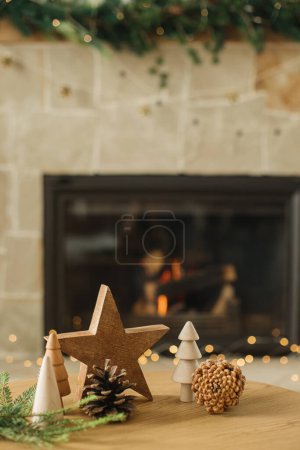 Téléchargez les photos : Joyeux Noël et bonnes vacances ! Élégants arbres de Noël en bois, étoiles, cônes de pin et branches de sapin sur la table contre la cheminée en feu. Décor moderne rustique respectueux de l'environnement dans le salon - en image libre de droit