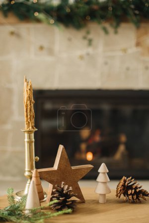 Foto de Feliz Navidad y felices fiestas! Elegantes árboles de madera de Navidad, estrellas, conos de pino y ramas de abeto en la mesa contra la chimenea en llamas. Moderna decoración ecológica rústica en sala de estar - Imagen libre de derechos