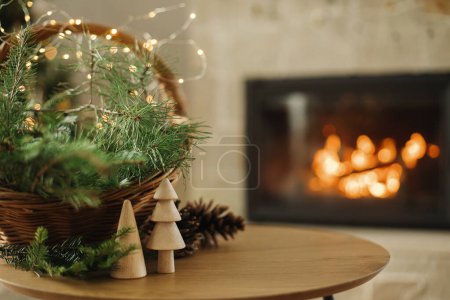 Photo for Merry Christmas and happy holidays! Stylish wicker basket with fir branches, wooden trees, pine cones on table against burning fireplace. Modern rustic eco friendly decor in living room - Royalty Free Image