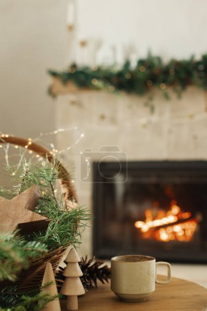 Photo for Cozy winter. Stylish cup of warm tea, basket with fir branches, wooden trees and star, pine cones on table against burning fireplace. Modern rustic eco friendly decor in scandinavian room - Royalty Free Image