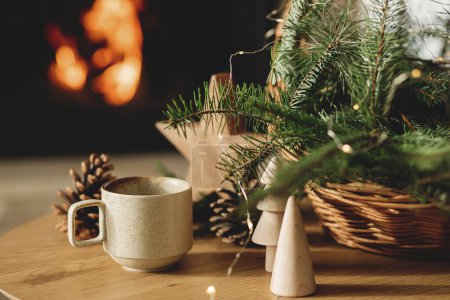 Photo for Cozy winter. Stylish cup of warm tea, basket with fir branches, wooden trees and star, pine cones on table against burning fireplace. Modern rustic eco friendly decor in scandinavian room - Royalty Free Image