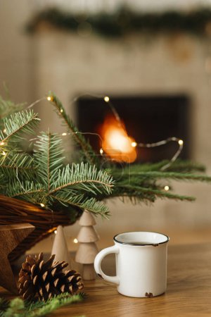 Photo for Stylish cup of warm tea, fir branches, wooden trees and star, pine cones on table against burning fireplace. Modern christmas rustic eco friendly decor and empty mug, scandinavian winter hygge - Royalty Free Image