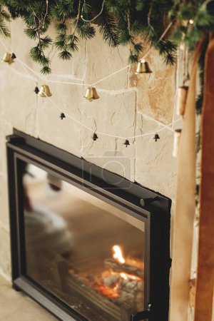 Foto de Elegante chimenea mantel decorado con ramas de Navidad, guirnalda campanas, adornos de madera. Decoración festiva rústica de la chimenea de Navidad en la moderna casa de campo sala de estar - Imagen libre de derechos