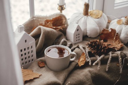 Foto de Autumn still life. Warm cup of tea, cozy pumpkin pillows, fall leaves, candle, lights and cute buildings decoration on brown scarf on windowsill. Hygge fall home decor. Happy Thanksgiving - Imagen libre de derechos