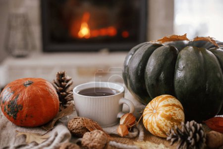 Foto de Warm cup of tea, pumpkins, autumn leaves, cozy scarf on rustic wooden table on background of burning fireplace. Hygge fall home, rural banner. Autumn still life. Happy Thanksgiving - Imagen libre de derechos