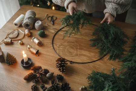 Téléchargez les photos : Je fais une couronne rustique de Noël. Mains tenant des branches de cèdre, faisant des couronnes sur une table en bois avec des cônes de pin, bougie, ficelle, cloches. Vue de dessus. Préparatifs des vacances d'hiver, temps atmosphérique - en image libre de droit