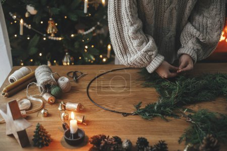 Téléchargez les photos : Je fais une couronne rustique de Noël. Mains tenant des branches de cèdre, faisant des couronnes sur une table en bois avec des cônes de pin, bougie, ficelle, cloches dans la salle de fête atmosphérique. Préparation des vacances d'hiver - en image libre de droit