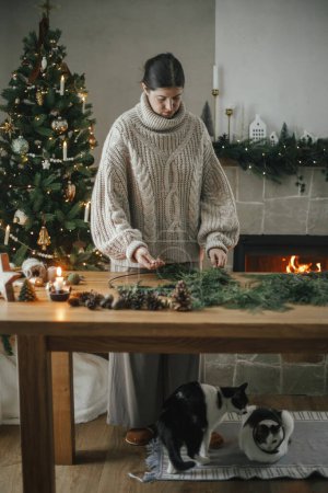 Téléchargez les photos : Femme en pull confortable faisant une couronne rustique de Noël sur une table en bois avec un chat mignon dans un salon festif avec cheminée. Préparatifs des vacances d'hiver, temps atmosphérique - en image libre de droit