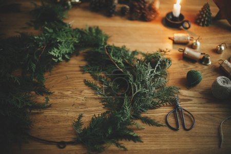 Photo for Making Christmas rustic wreath. Circle, cedar branches, pine cones, candle, twine, bells, scissors on wooden table in moody festive room. Winter holiday preparations, atmospheric time - Royalty Free Image