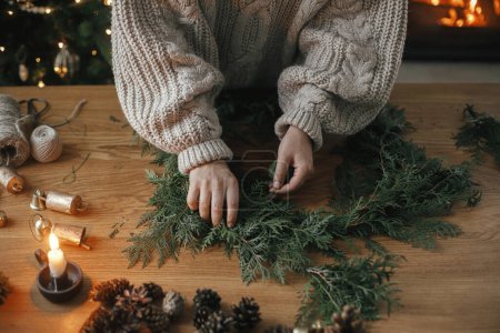 Photo for Making Christmas rustic wreath. Hands holding cedar branches, making wreath on wooden table with pine cones, candle, twine, bells in atmospheric festive room. Winter holiday preparations - Royalty Free Image