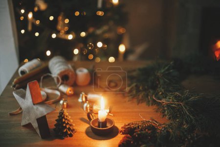 Téléchargez les photos : Bougie, branches de cèdre, cônes de pin, ficelle, cloches sur table en bois contre arbre et cheminée dans la salle du soir. Je fais une couronne rustique de Noël. Préparatifs des vacances d'hiver, temps atmosphérique - en image libre de droit