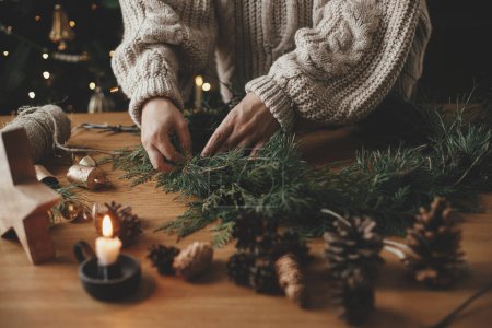 Photo for Hands in cozy sweater making Christmas rustic wreath with fir branches, pine cones, twine, bells on wooden table against festive tree, close up. Winter holiday preparations, atmospheric time - Royalty Free Image