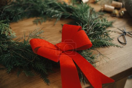 Foto de Corona con estilo de Navidad. Corona de Navidad con lazo rojo, ramas de cedro, tijeras en mesa de madera. Feliz Navidad. Preparativos para las vacaciones de invierno, tiempo atmosférico - Imagen libre de derechos