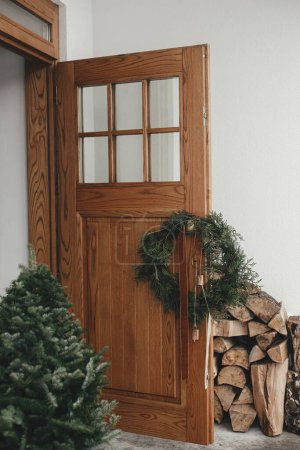 Foto de Elegante corona rústica de Navidad con campanas vintage y cinta colgando de mango de latón en la puerta delantera de madera y abeto. Moderna eco granja decoración exterior para vacaciones rurales de invierno - Imagen libre de derechos