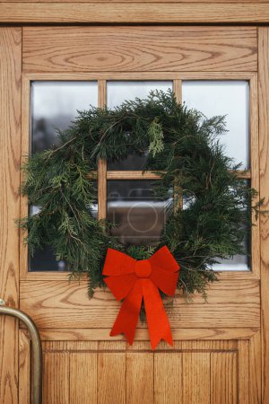 Photo for Stylish christmas rustic wreath with red bow on wooden doors at house entrance. Winter holiday decoration of modern farmhouse exterior. Merry Christmas and Happy holidays - Royalty Free Image