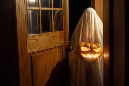 Téléchargez les photos : Fantôme effrayant avec Jack o lanterne rougeoyante aux portes dans la nuit atmosphérique effrayante. Joyeux Halloween ! Personne déguisée en fantôme ou traitant à l'entrée de la maison dans l'obscurité. - en image libre de droit