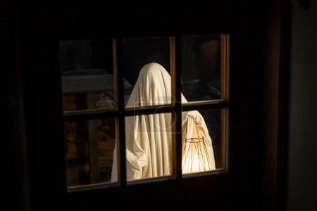 Téléchargez les photos : Fantôme effrayant avec lanterne lumineuse debout aux portes dans une nuit atmosphérique effrayante. Joyeux Halloween ! Personne déguisée en fantôme ou traitant à l'entrée de la maison dans l'obscurité. Temps d'horreur - en image libre de droit