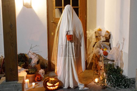 Téléchargez les photos : Trucs ou friandises ! Fantôme effrayant tenant un seau de bonbons d'Halloween sur le fond de l'entrée de la maison avec Jack o lanterne et effrayantes décorations d'Halloween atmosphériques dans l'obscurité. Joyeux Halloween - en image libre de droit