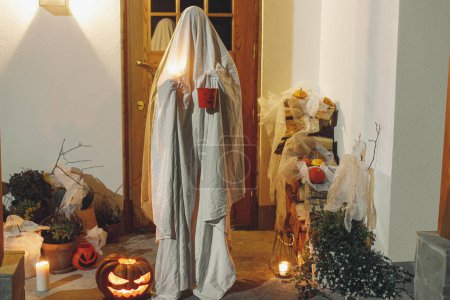 Foto de ¡Truco o trato! Fantasma espeluznante sosteniendo cubo de caramelo de Halloween en el fondo de la entrada de la casa con Jack o linterna y decoraciones de Halloween atmosféricas de miedo en la oscuridad. Feliz Halloween - Imagen libre de derechos